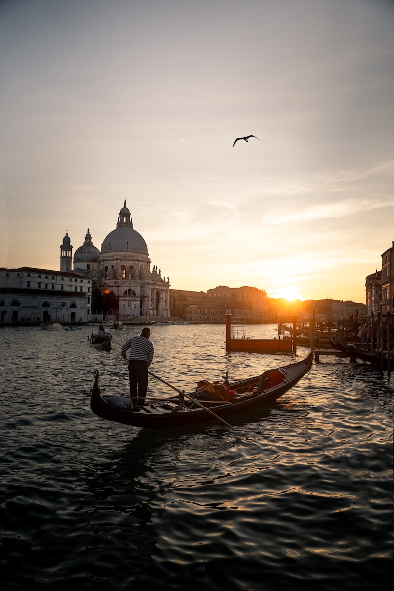 A traditional Venetian gondola glides gracefully along the calm waters of a canal, surrounded by historic pastel-colored buildings, while the gondolier in his distinctive attire steers the boat with skillful hands.