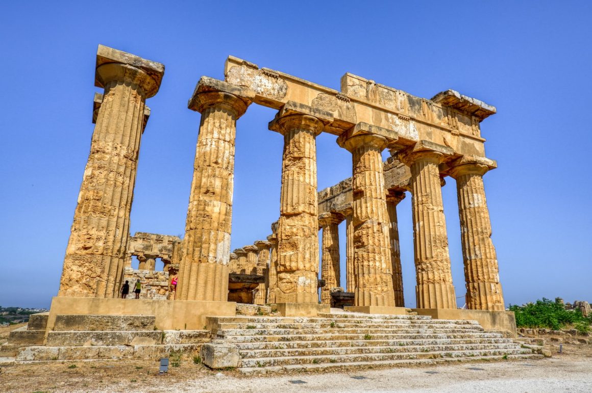 Panoramic view of the Valley of the Temples in Sicily, where ancient Greek temples rise against a clear blue sky, surrounded by a landscape of Mediterranean vegetation and olive trees.