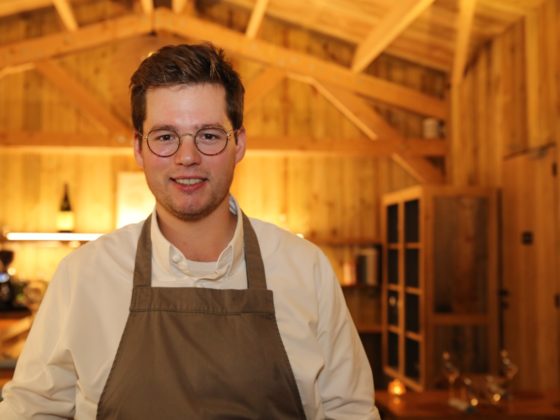 Chef Basile De Wulf in zijn restaurant, glimlachend en gekleed in een wit shirt en schort, met de rustieke houten binnenkant van de chalet op de achtergrond