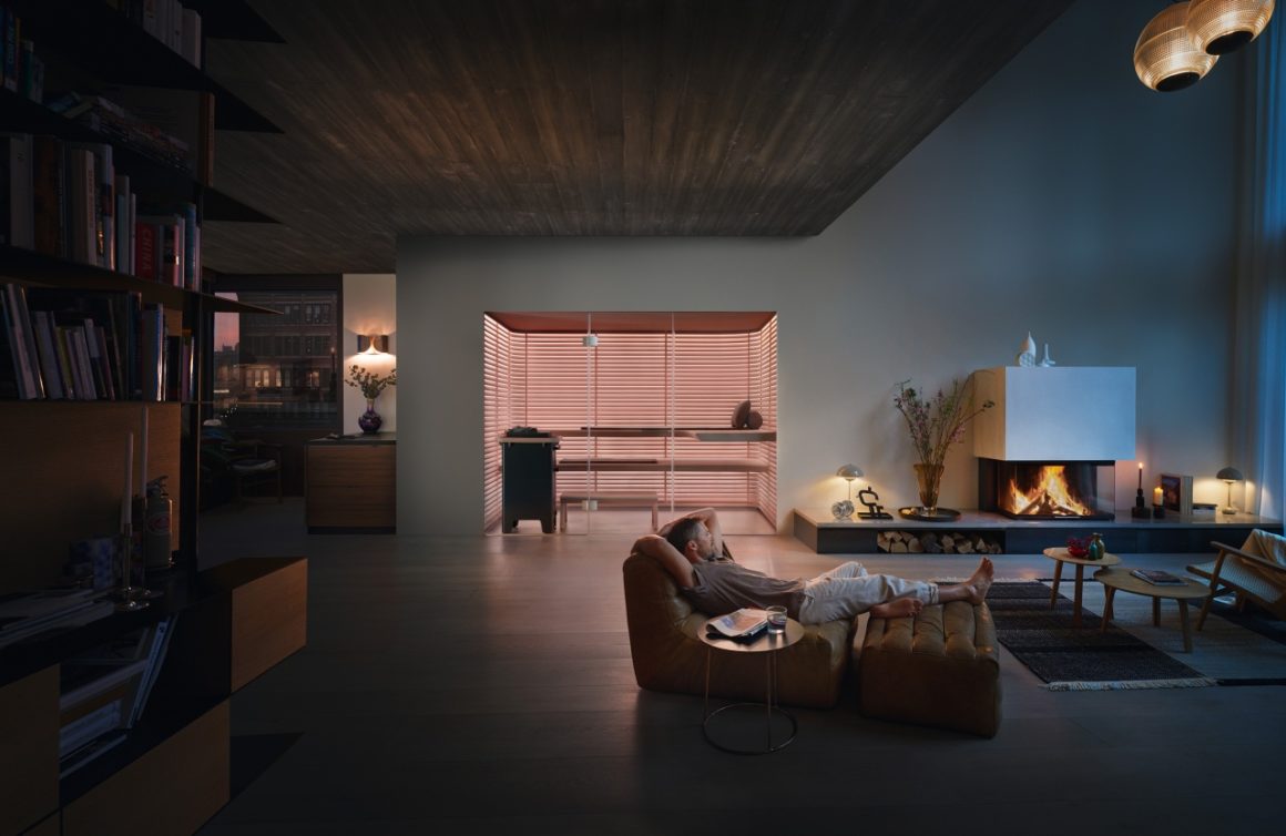  This image shows a spacious living room with an open design. On the left side, there is an integrated sauna with wooden slats and soft lighting. The living room itself has modern furniture, such as a comfortable sofa and a side table. Large windows offer panoramic views of the foreign landscape. There are also some glass pendant lights hanging, giving the interior a chic look.