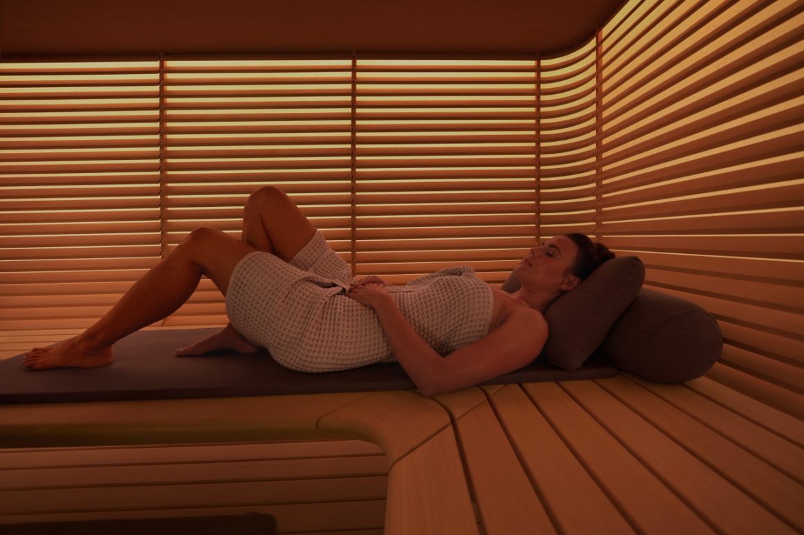 Het derde beeld laat een persoon zien die ontspant in de sauna. Het licht dat door de lamellen schijnt, geeft een warme gloed aan de ruimte