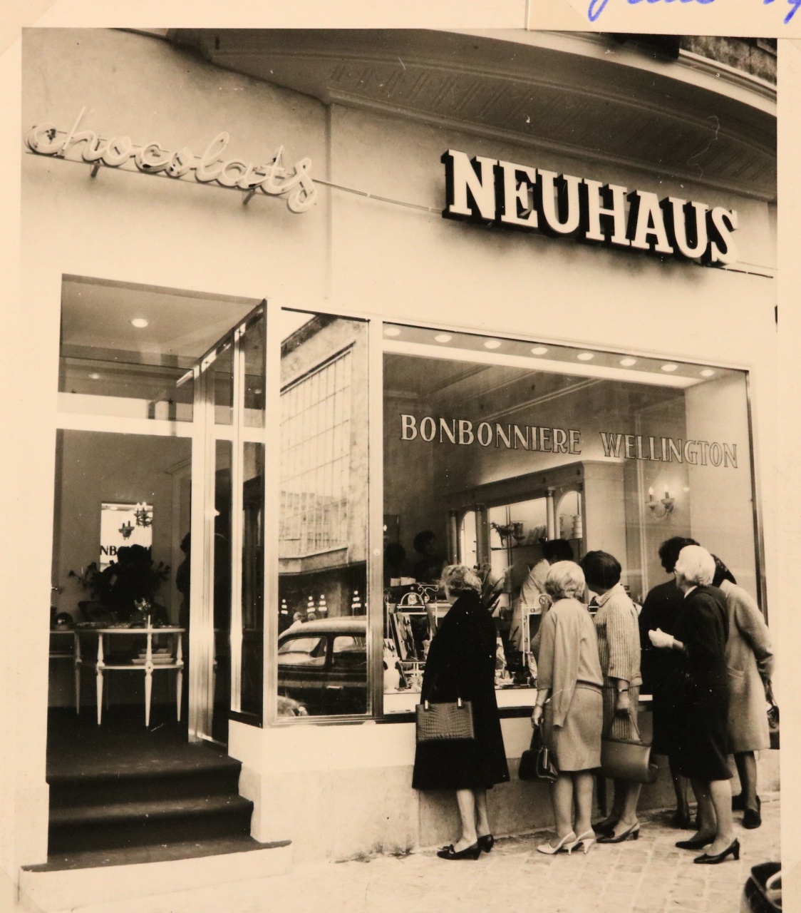Historische zwart-wit foto van de buitenkant van de Neuhaus chocolaterie, met klanten die voor de etalage staan