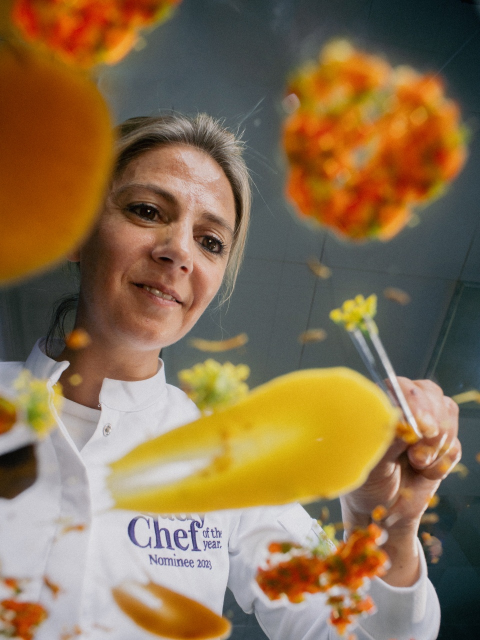 Een chef-kok die speels geel voedsel de lucht in gooit, met de tekst "Chef of the Year Nominee 2023" op haar jas