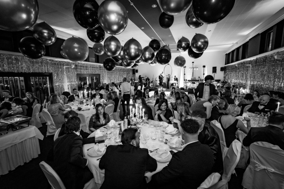 Een feestzaal met gasten aan ronde tafels onder grote zwarte en gouden ballonnen, met een band op de achtergrond.