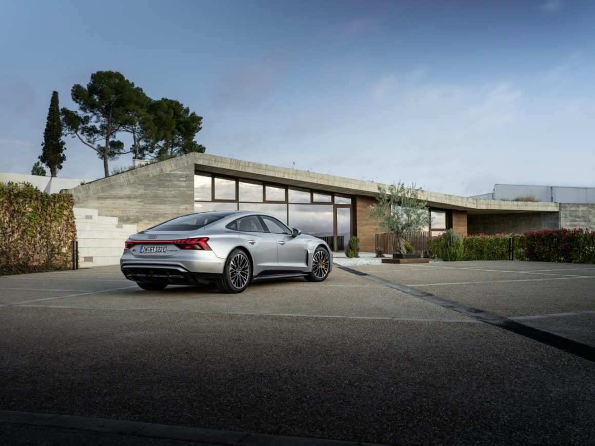 Audi e-tron GT geparkeerd voor een moderne woning met veel glas en beton, omgeven door groene beplanting. De auto staat stil en het landschap weerspiegelt een serene sfeer.