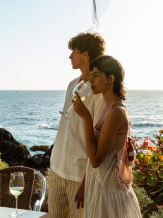 Een man en een vrouw genieten van een glas wijn bij een tafel aan de kust van Tenerife, met uitzicht op de oceaan en rotsachtige kliffen in de achtergrond. De zon schijnt helder, waardoor een serene en romantische sfeer ontstaat.