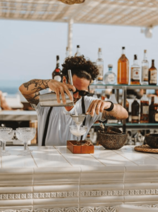 Mixologist aan het werk in een stijlvolle beachbar op Ibiza, giet een ambachtelijke cocktail door een fijne zeef in een glas, omringd door tropische sfeer, flessen sterke drank en de blauwe zee op de achtergrond.