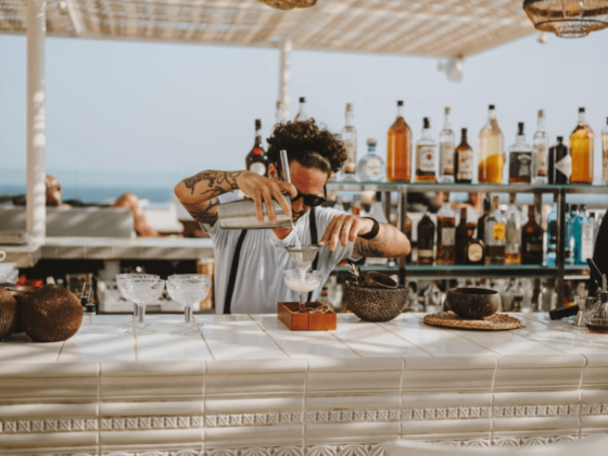 Mixologist aan het werk in een stijlvolle beachbar op Ibiza, giet een ambachtelijke cocktail door een fijne zeef in een glas, omringd door tropische sfeer, flessen sterke drank en de blauwe zee op de achtergrond.