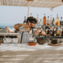 Mixologist aan het werk in een stijlvolle beachbar op Ibiza, giet een ambachtelijke cocktail door een fijne zeef in een glas, omringd door tropische sfeer, flessen sterke drank en de blauwe zee op de achtergrond.