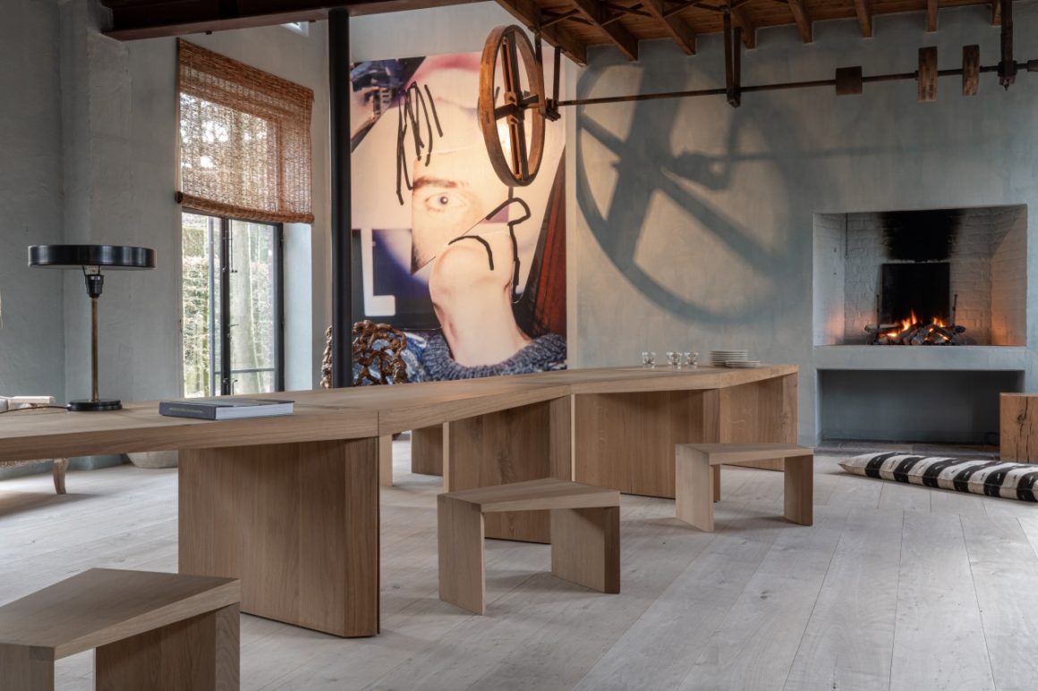 De Endless Table van Collected by Obumex in een karaktervolle ruimte met open haard, kunstzinnige muurdecoratie en houten balkenplafond. Het natuurlijke eikenhout van de tafel en bijhorende zitjes straalt vakmanschap uit. De zigzagvorm nodigt uit tot gesprekken en warme bijeenkomsten. Een sfeervolle setting waar design en authenticiteit samenkomen.