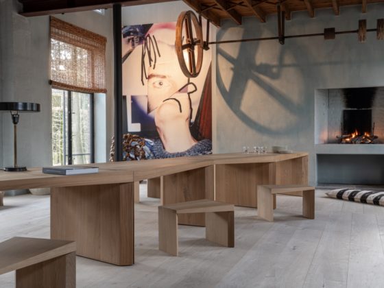 De Endless Table van Collected by Obumex in een karaktervolle ruimte met open haard, kunstzinnige muurdecoratie en houten balkenplafond. Het natuurlijke eikenhout van de tafel en bijhorende zitjes straalt vakmanschap uit. De zigzagvorm nodigt uit tot gesprekken en warme bijeenkomsten. Een sfeervolle setting waar design en authenticiteit samenkomen.