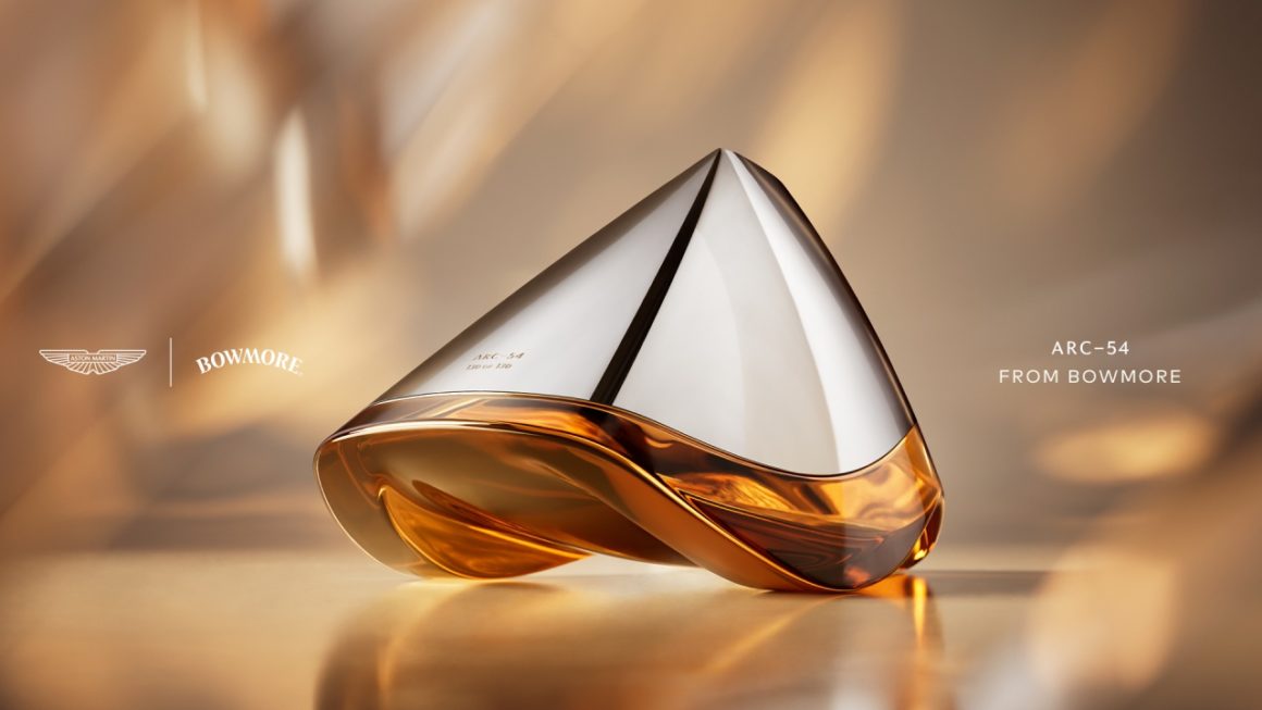 Close-up van de futuristische Bowmore ARC-54 whiskykaraf, ontworpen in samenwerking met Aston Martin. De handgeblazen decanter met zilveren top en amberkleurige whisky onderaan rust elegant op een warme, goudtintige achtergrond. Links staan de logo’s van Aston Martin en Bowmore, rechts de tekst “ARC-54 from Bowmore
