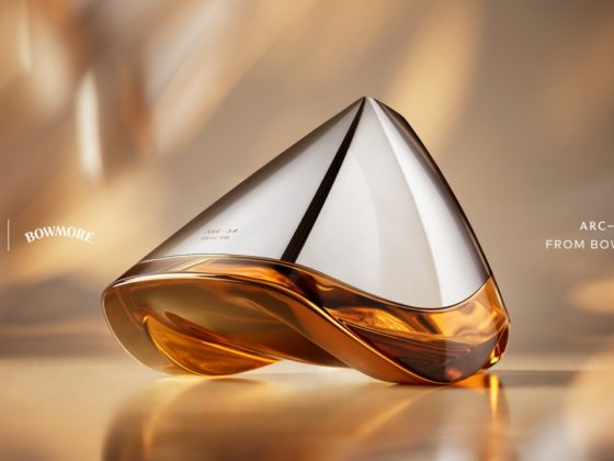 Close-up van de futuristische Bowmore ARC-54 whiskykaraf, ontworpen in samenwerking met Aston Martin. De handgeblazen decanter met zilveren top en amberkleurige whisky onderaan rust elegant op een warme, goudtintige achtergrond. Links staan de logo’s van Aston Martin en Bowmore, rechts de tekst “ARC-54 from Bowmore