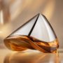 Close-up van de futuristische Bowmore ARC-54 whiskykaraf, ontworpen in samenwerking met Aston Martin. De handgeblazen decanter met zilveren top en amberkleurige whisky onderaan rust elegant op een warme, goudtintige achtergrond. Links staan de logo’s van Aston Martin en Bowmore, rechts de tekst “ARC-54 from Bowmore