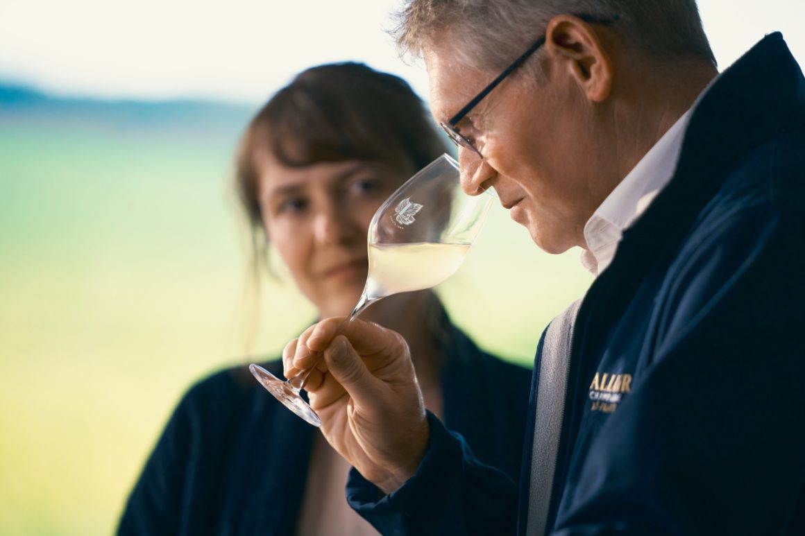 Keldermeester Dominique Demarville van Champagne Lallier ruikt aandachtig aan een glas champagne tijdens een proeverij, terwijl een vrouwelijke collega toekijkt in een natuurlijke setting met zacht groen op de achtergrond.
