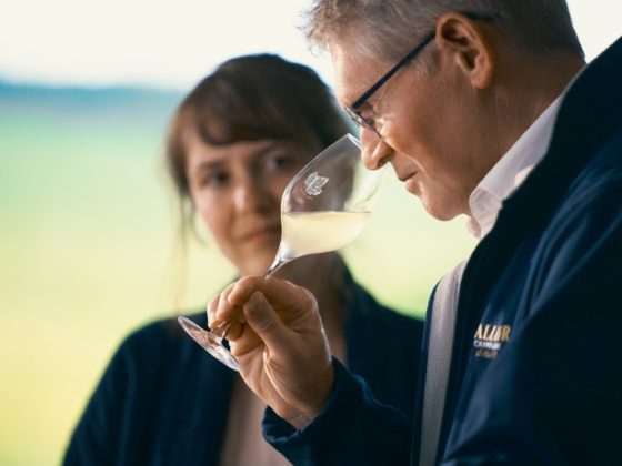 Keldermeester Dominique Demarville van Champagne Lallier ruikt aandachtig aan een glas champagne tijdens een proeverij, terwijl een vrouwelijke collega toekijkt in een natuurlijke setting met zacht groen op de achtergrond.