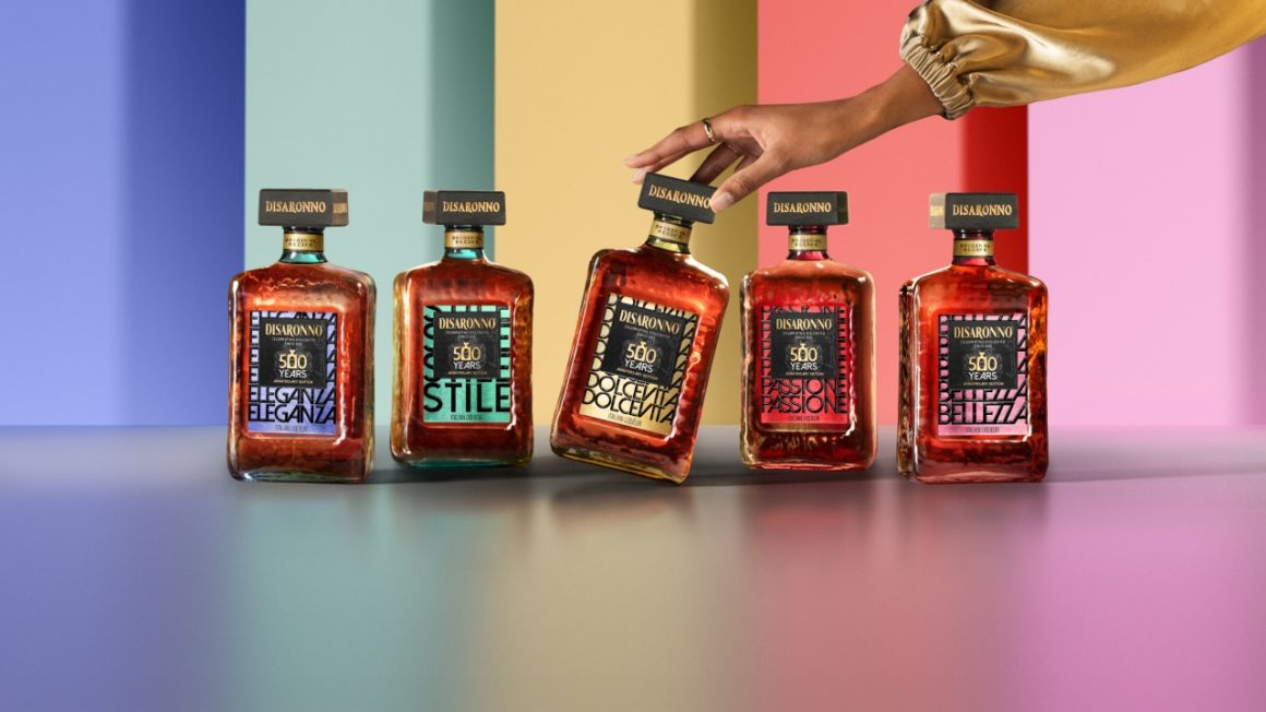 De vijf Disaronno 500 jaar limited edition flessen in een kleurrijke setting, waarbij een hand de Dolce Vita-fles elegant aanraakt. English: The five Disaronno 500-year limited edition bottles displayed in a vibrant setting, with a hand gently touching the Dolce Vita bottle.