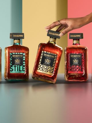 De vijf Disaronno 500 jaar limited edition flessen in een kleurrijke setting, waarbij een hand de Dolce Vita-fles elegant aanraakt. English: The five Disaronno 500-year limited edition bottles displayed in a vibrant setting, with a hand gently touching the Dolce Vita bottle.