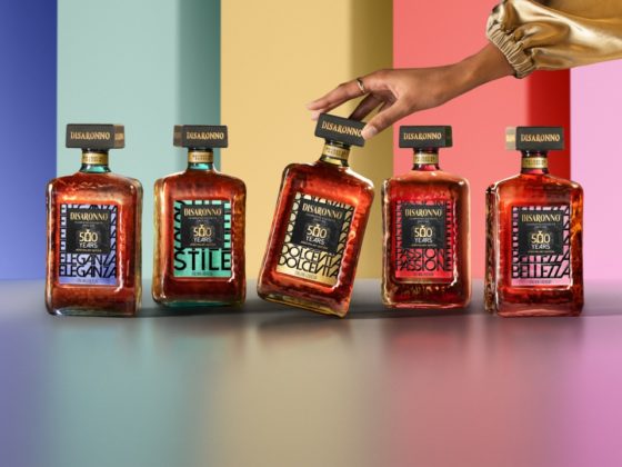 De vijf Disaronno 500 jaar limited edition flessen in een kleurrijke setting, waarbij een hand de Dolce Vita-fles elegant aanraakt. English: The five Disaronno 500-year limited edition bottles displayed in a vibrant setting, with a hand gently touching the Dolce Vita bottle.