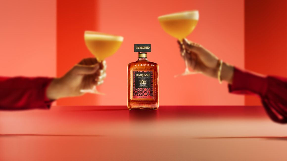 Disaronno Passione limited edition fles centraal in beeld met twee handen die toosten met elegante cocktails in een warme rode setting.